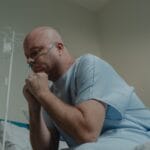 Łysy pacjent w szpitalnej koszuli siedzi zamyślony z rurką tlenową